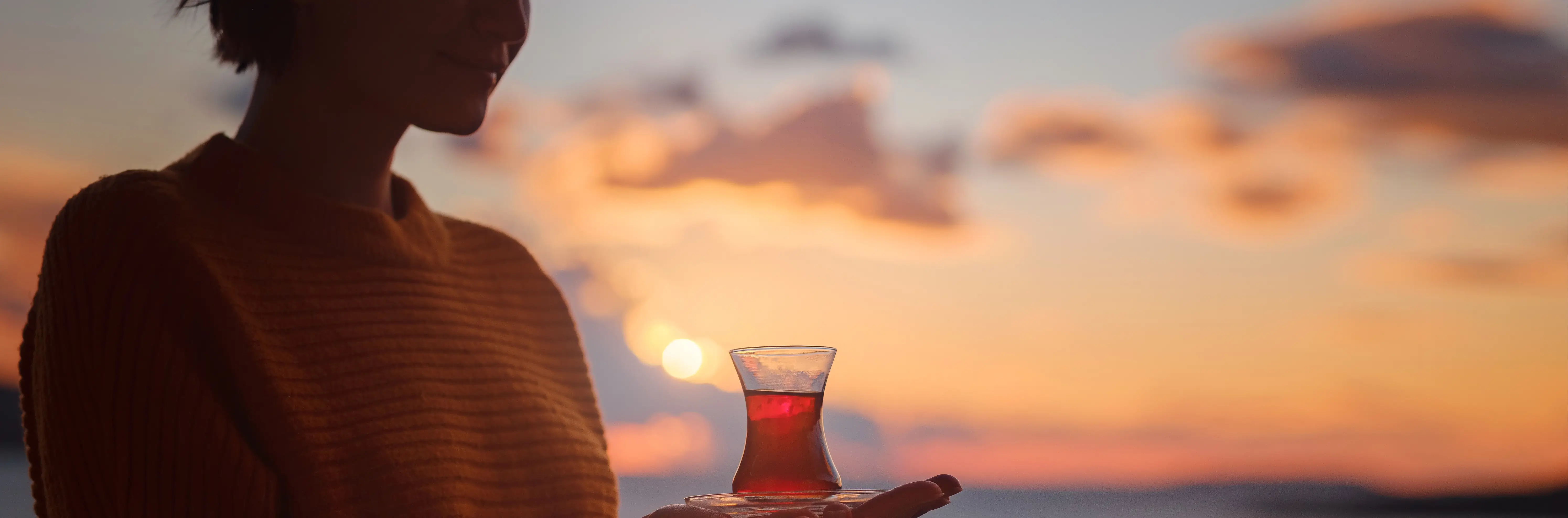 A person in silhouette holding a glass of tea against a warm sunset over water